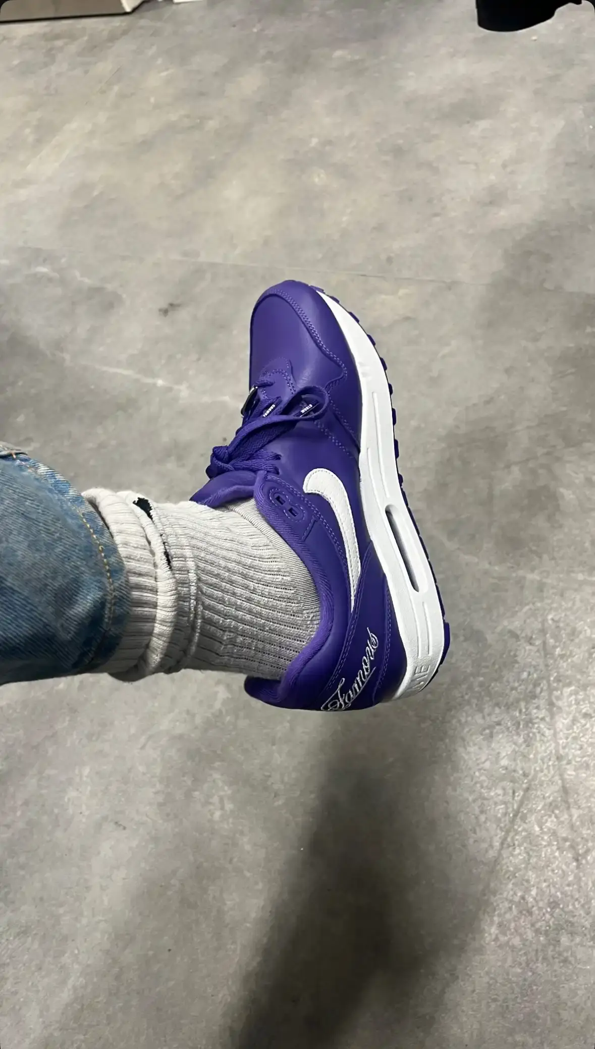 @_clementowusu Close-up photograph of the Supreme x Nike Air Max 1 '87 in Varsity Purple colorway showing white Supreme "World Famous" script on the purple heel counter, white midsole with debossed SUPREME wordmark, and purple upper with white Nike swoosh logo on the side.