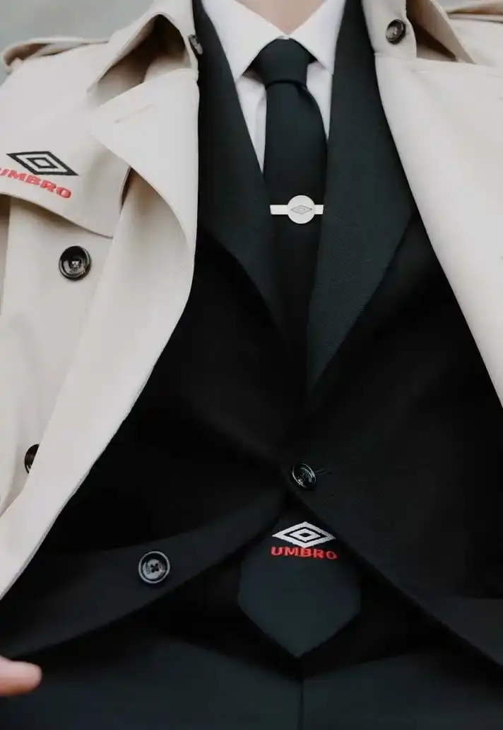 UMBRO AND LC23 REVIVE FOOTBALL'S FORMAL ERA photo of fashion collaboration between sportswear brand Umbro and Italian label LC23 for Spring/Summer 2025. The collection draws inspiration from 1990s football manager attire, featuring items such as a leather Speciali sneaker with interchangeable tongue, a beige trench coat, a knitted football shirt, and accessories including a blazer, shorts, tie, and cap. The collection blends formal touchline style with contemporary streetwear and will be available from April 23 on lc23.it.