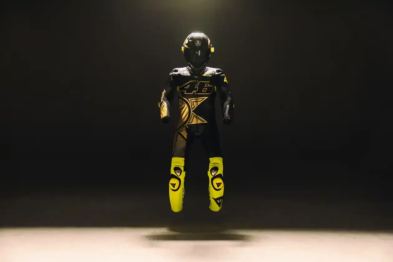 Dainese's Soleluna Vale46 Anniversary Kit displaying a black racing suit and helmet with yellow accents and gold leaf details, positioned in a glass display case. The gear features Valentino Rossi's signature number 46.