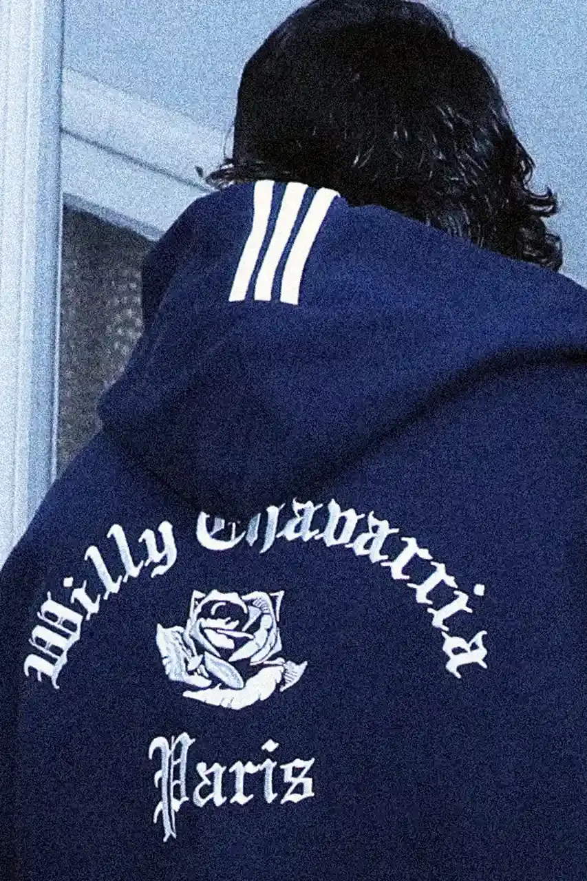 adidas Originals/Willy Chavarria Image shows a vintage Chevrolet Impala parked on a Parisian street, displaying Willy Chavarria x adidas Originals limited-edition apparel and footwear collection, featuring navy t-shirts, hoodies, and sneakers with rose motifs and collaborative branding. (image 3 of 7)