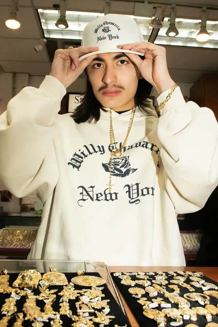 Willy Chavarria Image of Willy Chavarria's New York Collection featuring clothing items with Old English typography and the brand's signature rose logo. The collection includes hoodies, t-shirts, and caps in navy blue and off-white colors that showcase 'Willy Chavarria' arched above a rose emblem with 'New York' text below. (image 3 of 5)