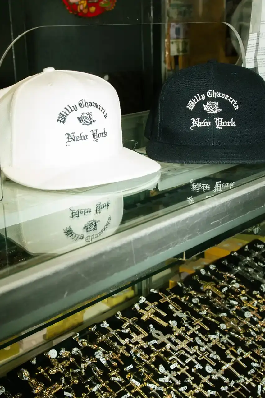 Willy Chavarria Image of Willy Chavarria's New York Collection featuring clothing items with Old English typography and the brand's signature rose logo. The collection includes hoodies, t-shirts, and caps in navy blue and off-white colors that showcase 'Willy Chavarria' arched above a rose emblem with 'New York' text below. (image 5 of 5)