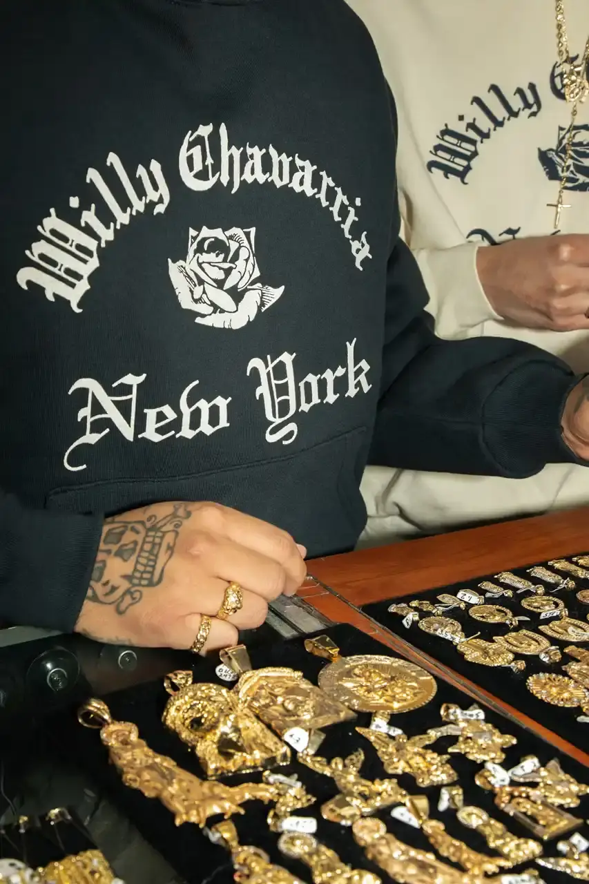 Willy Chavarria Image of Willy Chavarria's New York Collection featuring clothing items with Old English typography and the brand's signature rose logo. The collection includes hoodies, t-shirts, and caps in navy blue and off-white colors that showcase 'Willy Chavarria' arched above a rose emblem with 'New York' text below. (image 2 of 5)