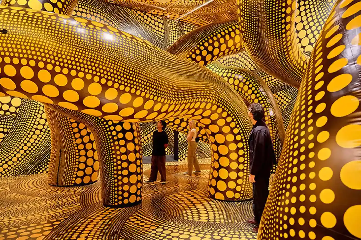 Sean Fennessy/Yayoi Kusama A photograph of Yayoi Kusama's immersive infinity mirrored room, featuring reflective surfaces creating a kaleidoscopic environment of endless light and geometric patterns. Polka-dotted sculptures and vibrant yellow and black elements are visible, representing the artist's signature visual style.