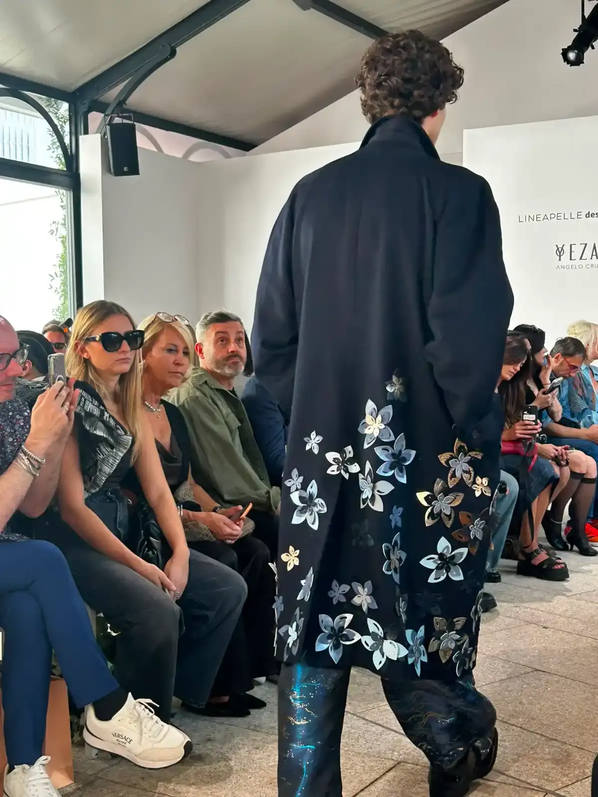 YEZAEL "VERITÀ" COLLECTION BY ANGELO CRUCIANI image of Marche-born designer Angelo Cruciani's "Verità" collection for Yezael presented at Fashion Week 2026. The text describes tailored suits with wide-leg trousers, 3D floral appliqués, an ochre-yellow coat, a pastel-blue Rococo-inspired suit, and contrasting textures combining leather and pearls. Key quotes and philosophical reflections explore fashion, consumerism, nature, and personal identity. (image 12 of 12)
