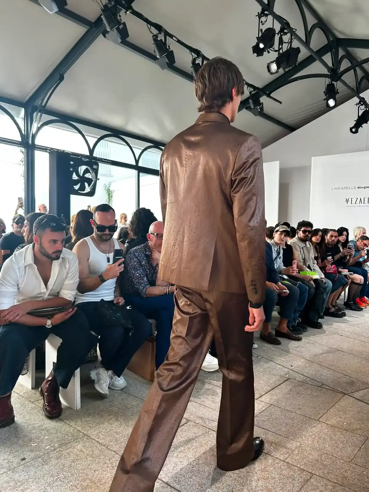 YEZAEL "VERITÀ" COLLECTION BY ANGELO CRUCIANI image of Marche-born designer Angelo Cruciani's "Verità" collection for Yezael presented at Fashion Week 2026. The text describes tailored suits with wide-leg trousers, 3D floral appliqués, an ochre-yellow coat, a pastel-blue Rococo-inspired suit, and contrasting textures combining leather and pearls. Key quotes and philosophical reflections explore fashion, consumerism, nature, and personal identity. (image 3 of 12)