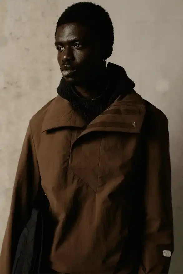 Samuel Ross Models on a darkened runway at Paris Fashion Week wearing Samuel Ross's ZARA collection. The pieces include structured outerwear with cinched waists, white cargo shorts, and minimalist coats. The presentation features digital projections in the background, creating a dramatic atmosphere as models showcase the FW25 collection's muted color palette and utility-focused designs. (image 4 of 12)