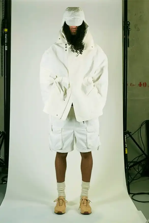 Samuel Ross Models on a darkened runway at Paris Fashion Week wearing Samuel Ross's ZARA collection. The pieces include structured outerwear with cinched waists, white cargo shorts, and minimalist coats. The presentation features digital projections in the background, creating a dramatic atmosphere as models showcase the FW25 collection's muted color palette and utility-focused designs.