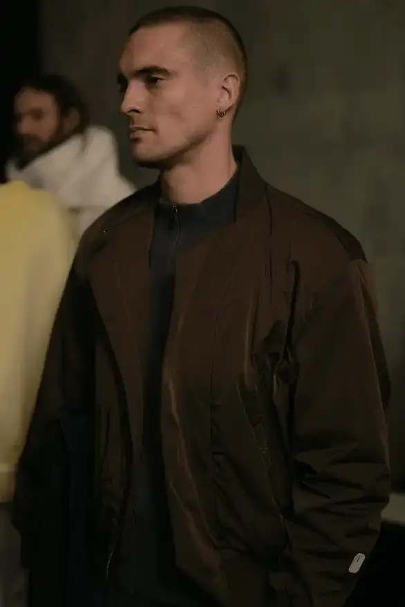 Samuel Ross Models on a darkened runway at Paris Fashion Week wearing Samuel Ross's ZARA collection. The pieces include structured outerwear with cinched waists, white cargo shorts, and minimalist coats. The presentation features digital projections in the background, creating a dramatic atmosphere as models showcase the FW25 collection's muted color palette and utility-focused designs. (image 6 of 12)