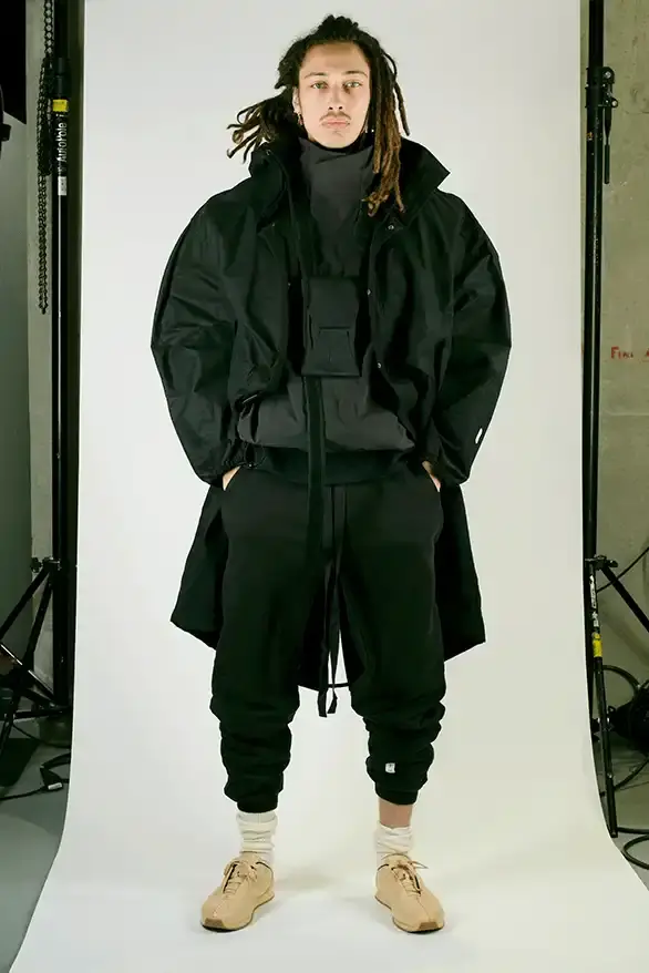 Samuel Ross Models on a darkened runway at Paris Fashion Week wearing Samuel Ross's ZARA collection. The pieces include structured outerwear with cinched waists, white cargo shorts, and minimalist coats. The presentation features digital projections in the background, creating a dramatic atmosphere as models showcase the FW25 collection's muted color palette and utility-focused designs. (image 5 of 12)