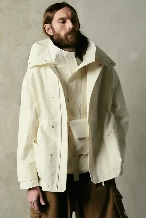 Samuel Ross Models on a darkened runway at Paris Fashion Week wearing Samuel Ross's ZARA collection. The pieces include structured outerwear with cinched waists, white cargo shorts, and minimalist coats. The presentation features digital projections in the background, creating a dramatic atmosphere as models showcase the FW25 collection's muted color palette and utility-focused designs. (image 3 of 12)