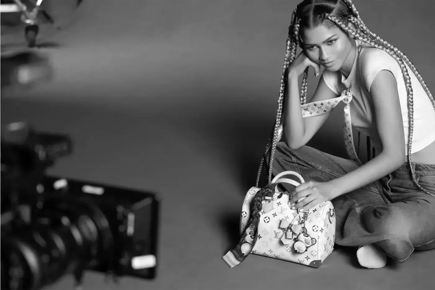 Louis Vuitton Zendaya poses in casual denim and a white crop top against a green screen background with Murakami's floral motifs. She wears a white Louis Vuitton x Murakami bandeau and holds a matching Keepall bag (image 5 of 5)